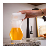 Hand pouring beer from a pitcher into a glass on a kitchen counter.